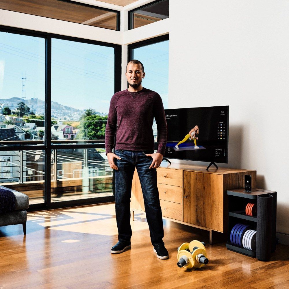 Moawia Eldeeb stands in a bright apartment beside a TV showing a Tempo class, with Tempo weights and their storage next to him.